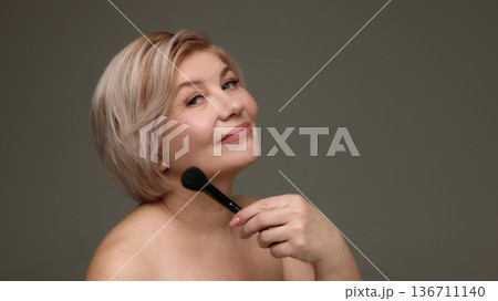 Mature woman posing with makeup brush near chin. Mature woman posing with makeup brush near chin. 136711140