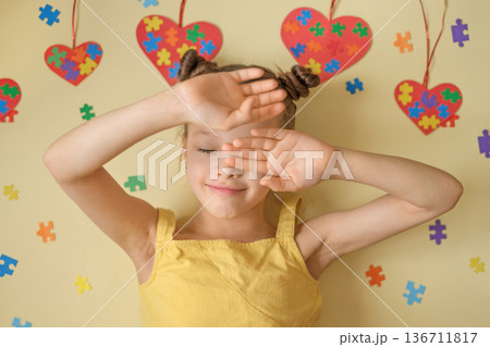 Shy girl covering face with hands against yellow background decorated with colorful puzzle. Concept of autism awareness, sensory sensitivity, introversion, emotional vulnerability and support for kids Shy girl covering face with hands against yellow background decorated with colorful puzzle. Concept of autism awareness, sensory sensitivity, introversion, emotional vulnerability and support for kids 136711817