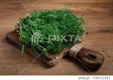 Freshly harvested pea shoots displayed on a rustic wooden cutting board against a textured wooden backdrop Freshly harvested pea shoots displayed on a rustic wooden cutting board against a textured wooden backdrop 136711931