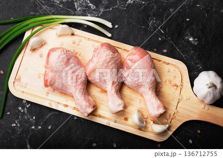 Top view of three raw chicken drumsticks on a wooden cutting board with garlic cloves, green onions, and pink salt, set against a dark marble background. High quality photo 136712755