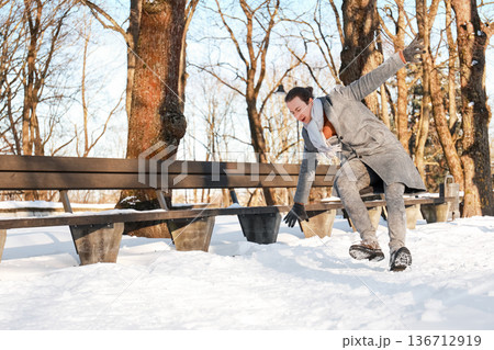 Man slipping and falling on snow near a bench in a winter park. Young guy losing balance on slippery ice during a sunny cold day. Winter accident and safety concept. High quality photo Man slipping and falling on snow near a bench in a winter park. Young guy losing balance on slippery ice during a sunny cold day. Winter accident and safety concept. High quality photo 136712919