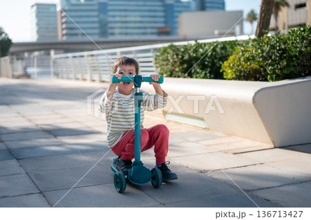 Young Boy Riding A Teal Scooter In Sunny Abu Dhabi Outdoor Scene 136713427