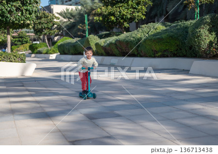 Young Boy Riding A Scooter In Playful Outdoor Setting With Joy. 136713438