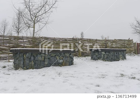 Snow-covered benches in a nature park. Benches coated in snow with rustic wooden fencing in the background. Concept of winter and winter nature. Snow-covered benches in a nature park. Benches coated in snow with rustic wooden fencing in the background. Concept of winter and winter nature. 136713499