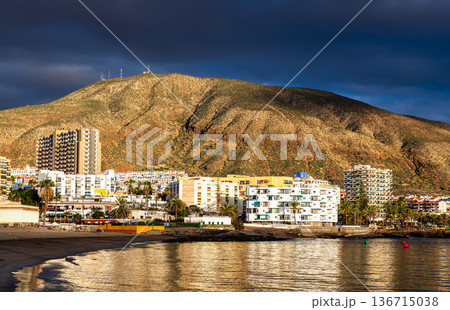 View of Playa de Los Cristianos beach and Mount Guaza in Tenerife, Canary Islands, Spain. Features the sandy beach, calm ocean water, and resort buildings illuminated at sunset. View of Playa de Los Cristianos beach and Mount Guaza in Tenerife, Canary Islands, Spain. Features the sandy beach, calm ocean water, and resort buildings illuminated at sunset. 136715038