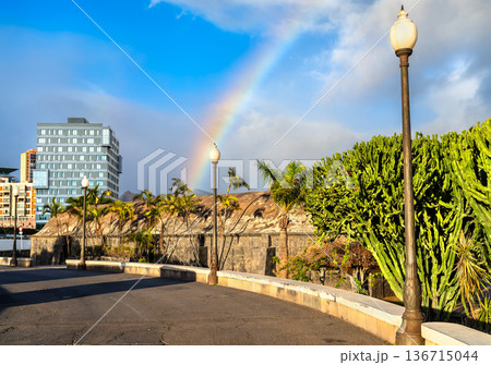 Rainbow over the historic Casa de la Polvora and modern buildings in Santa Cruz de Tenerife, Canary Islands, Spain. Features the stone walls of the 18th-century gunpowder house and a paved walkway. 136715044