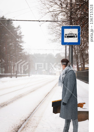 Man in a grey coat stands at a snowy tram stop, looking down the empty tracks. He waits impatiently for public transport on a cold winter day, fearing he is late. High quality photo Man in a grey coat stands at a snowy tram stop, looking down the empty tracks. He waits impatiently for public transport on a cold winter day, fearing he is late. High quality photo 136715328