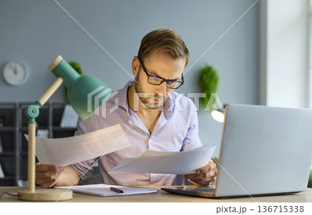 Serious accountant reviewing financial documents at laptop during audit work 136715338