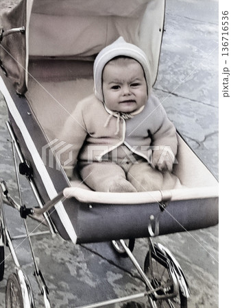A vintage portrait of a little boy sitting in a stroller. The baby is wearing a bonnet and cotton gloves. Retro photo from 1968. 136716536