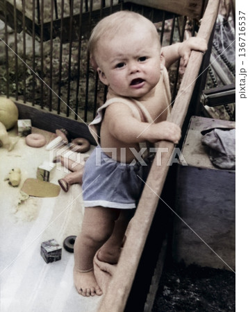 A vintage portrait of a little boy standing in a wooden playpen surrounded by toys. Retro photo from 1968. A vintage portrait of a little boy standing in a wooden playpen surrounded by toys. Retro photo from 1968. 136716537