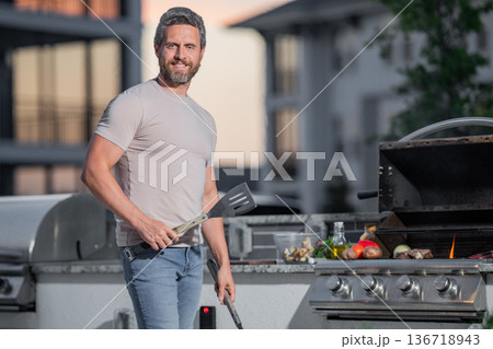 Handsome man preparing barbecue. Male cook cooking meat on barbecue grill. Guy cooking meat on barbecue for summer family dinner at the backyard of the house. 136718943
