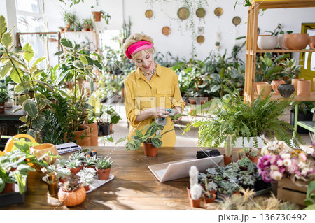 A woman tends to plants in a bright, airy greenhouse, surrounded by lush greenery and flowers 136730492