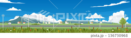 Landscape of an empty asphalt road. Cutting through wide fields lined with power poles. Small village sits on hill with meadows and flowers in foreground. Mountains in the background under a blue sky. 136730968