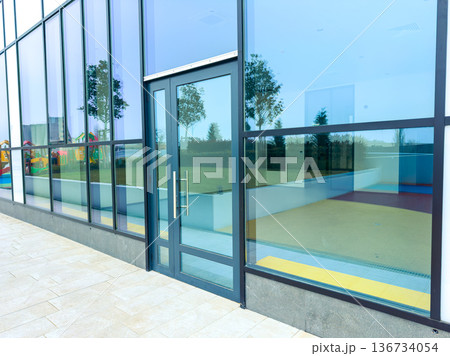 Building with large glass walls has a door and shows playground area in the background during bright day Building with large glass walls has a door and shows playground area in the background during bright day 136734054