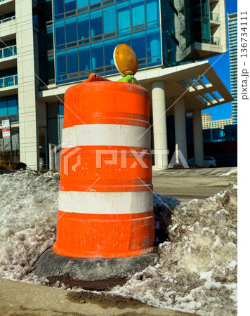 Construction cone stands on snow near modern building in city during winter day with clear blue sky above 136734111