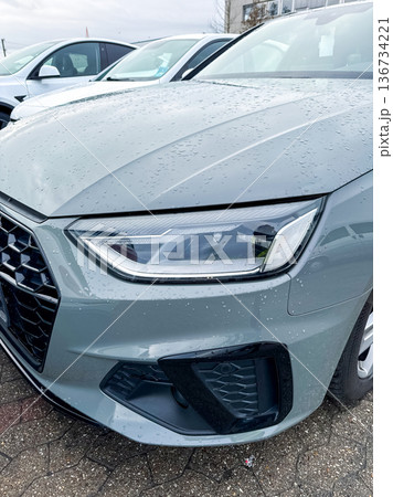 Close up view of a grey car parked among other vehicles at a dealership on a cloudy day with rain on the surface 136734221
