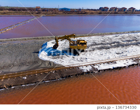 Excavator working on salt extraction near a pink lagoon through a sand path beside railway tracks 136734330