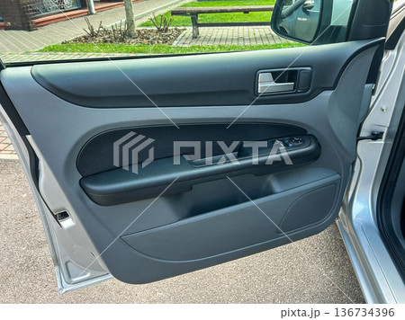 Interior view of a car door showing handle and controls. The door is open and outside is a parking area with trees and grass 136734396