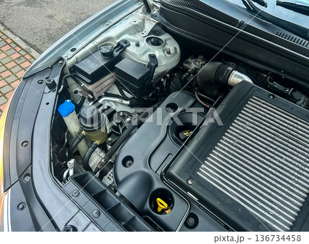 Engine compartment shows various components in a silver car with a visible air filter and fluid reservoir on a street during day time 136734458