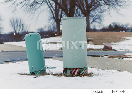Two green utility poles stand on the edge of a snow-covered area near a sidewalk in a suburban park during winter Two green utility poles stand on the edge of a snow-covered area near a sidewalk in a suburban park during winter 136734463