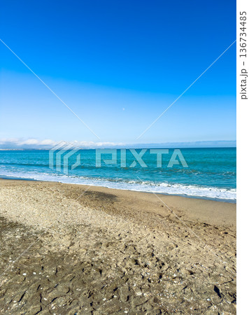 Bright day at the beach by the ocean with clear blue sky and gentle waves rolling onto the sandy shore under the warm sunlight during midday 136734485