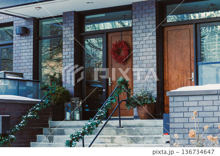 Decorative entrance in winter with dark bricks, wooden doors, wreath, and green garland near a snowy outdoor setting Decorative entrance in winter with dark bricks, wooden doors, wreath, and green garland near a snowy outdoor setting 136734647