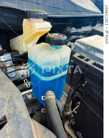 Coolant reservoir is seen in a vehicle engine bay showing blue fluid and other components nearby during daylight 136734687