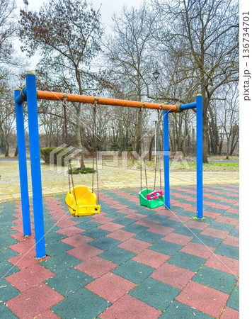 Swings are placed at a playground surrounded by trees on a cloudy day in the afternoon in a park setting 136734701
