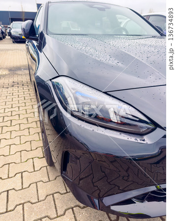Close view of a dark car parked in a lot with wet surface after rain, showing details of the headlight and body design, taken during a cloudy day Close view of a dark car parked in a lot with wet surface after rain, showing details of the headlight and body design, taken during a cloudy day 136734893