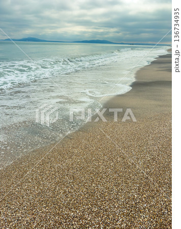 Waves crash onto a sandy beach during cloudy weather near a calm ocean shoreline in the early morning 136734935