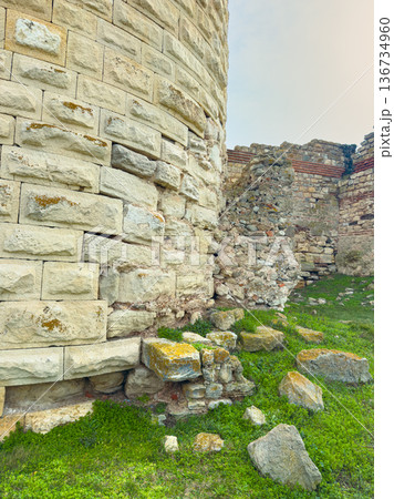 Ancient stone wall stands in ruins at historical site surrounded by grass and rocks in bright daylight 136734960