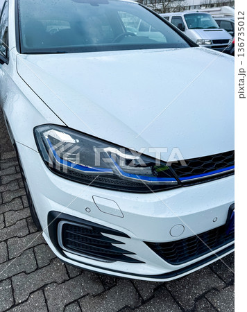 New car parked in a lot with wet pavement and cloudy weather showing details of the headlight and front grille while no other vehicles are nearby 136735012