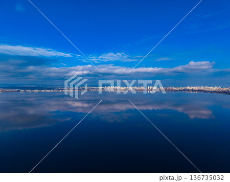 Calm water reflects blue sky and city skyline during midday at the lakeshore Calm water reflects blue sky and city skyline during midday at the lakeshore 136735032