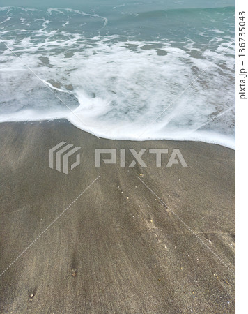 Waves wash over the sandy beach during late morning hours near the shoreline, creating patterns in the sand and foam as water retreats back into the ocean Waves wash over the sandy beach during late morning hours near the shoreline, creating patterns in the sand and foam as water retreats back into the ocean 136735043