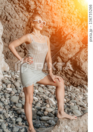 Woman beach rock posing on a pebble shore near cliffs during golden hour with white sunglasses Woman beach rock posing on a pebble shore near cliffs during golden hour with white sunglasses 136735266
