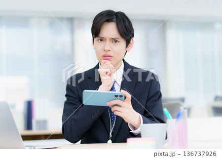 オフィスでスマホで動画視聴する困った顔の若い日本人男性 オフィスでスマホで動画視聴する困った顔の若い日本人男性 136737926