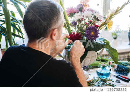Rear view of man carefully arranging aster flowers in stone urn. Men's mental health, creative therapy, mindful crafting, emotional regulation, stress relief for men, breaking stereotypes. Rear view of man carefully arranging aster flowers in stone urn. Men's mental health, creative therapy, mindful crafting, emotional regulation, stress relief for men, breaking stereotypes. 136738823
