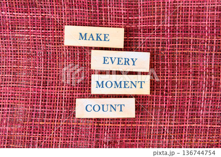 Four wooden blocks spell MAKE EVERY MOMENT COUNT on a rich crimson woven background, offering a daily dose of wisdom 136744754