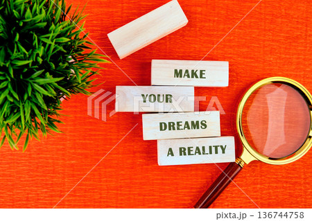 MAKE YOUR DREAMS A REALITY in wooden blocks on orange, with green foliage and a golden magnifying glass 136744758