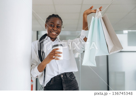 Smiling, holding shopping bags and drink. Businesswoman in formal clothes is in the modern room 136745348