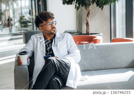 Holding paper cup of drink, break. Doctor is indoors in the modern building 136746255