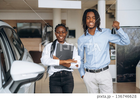 Happy owner is holding keys. Car seller is assisting client in the automobile salon Happy owner is holding keys. Car seller is assisting client in the automobile salon 136746628
