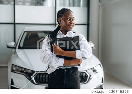 Black notepad in hands, smiling. Car seller, businesswoman is standing in automobile salon 136746898