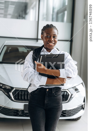 Black notepad in hands, smiling. Car seller, businesswoman is standing in automobile salon 136746899