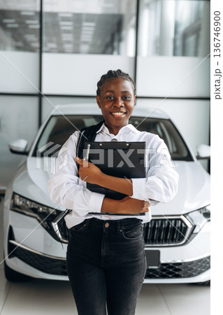 Black notepad in hands, smiling. Car seller, businesswoman is standing in automobile salon Black notepad in hands, smiling. Car seller, businesswoman is standing in automobile salon 136746900