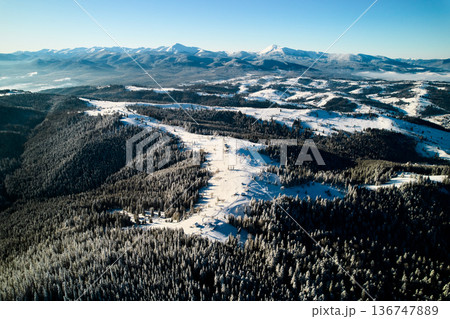 Aerial view of snow-covered valley with winding road passing through small village. Landscape features gently rolling hills, dense forests, and distant mountains under clear blue sky, with soft mist. Aerial view of snow-covered valley with winding road passing through small village. Landscape features gently rolling hills, dense forests, and distant mountains under clear blue sky, with soft mist. 136747889