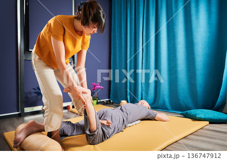 Masseuse making thai yoga massage. Woman therapist doing traditional massage treatment, stretching male patient's body. Client lying on mat, enjoying hand massage in calm atmosphere. Masseuse making thai yoga massage. Woman therapist doing traditional massage treatment, stretching male patient's body. Client lying on mat, enjoying hand massage in calm atmosphere. 136747912
