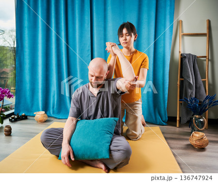 Masseuse making thai yoga massage. Woman therapist doing traditional massage treatment for male patient indoors. Bald client sitting on mat, meditating, enjoying hand massage in calm atmosphere. 136747924