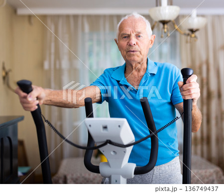 Elderly man exercising on elliptic trainer in living room Elderly man exercising on elliptic trainer in living room 136749437
