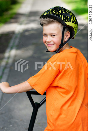 A young boy with blond hair and an orange T-shirt on a scooter 136750133